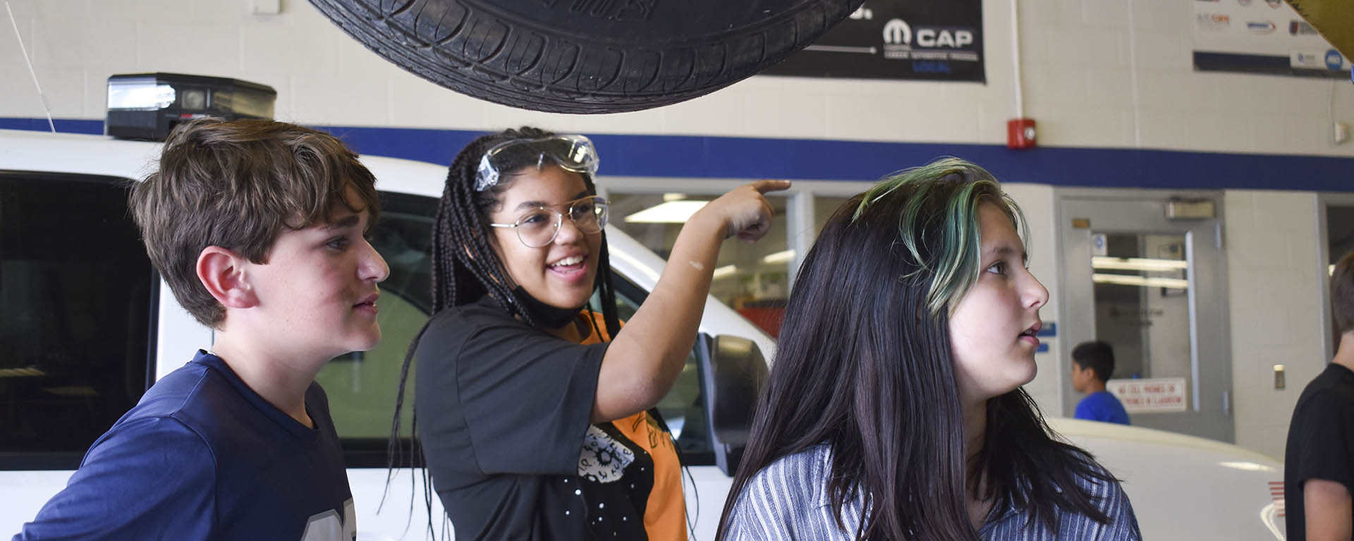 A student smiles and points while a car is suspended in the shop overhead.